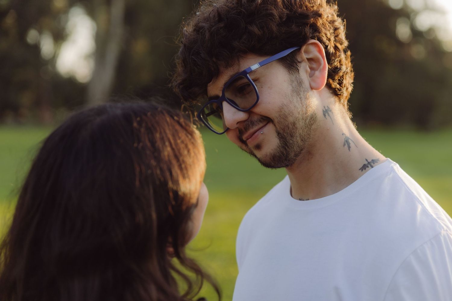 Young couple sharing a romantic moment outdoors in a sunny park, man with curly hair and glasses smiling warmly.