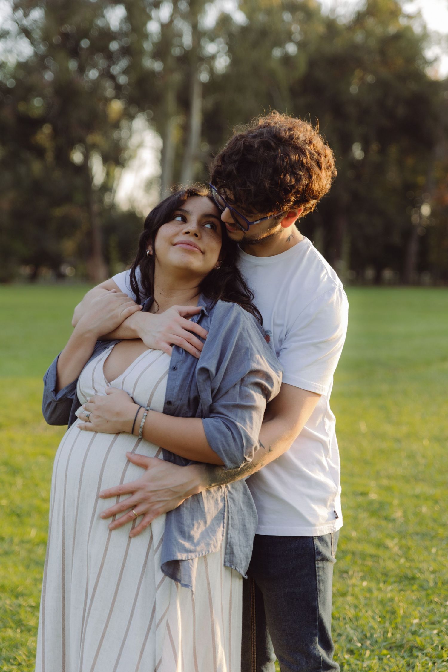 Tattooed man kissing pregnant woman's head from behind in a sunny park, both smiling in warm golden light.