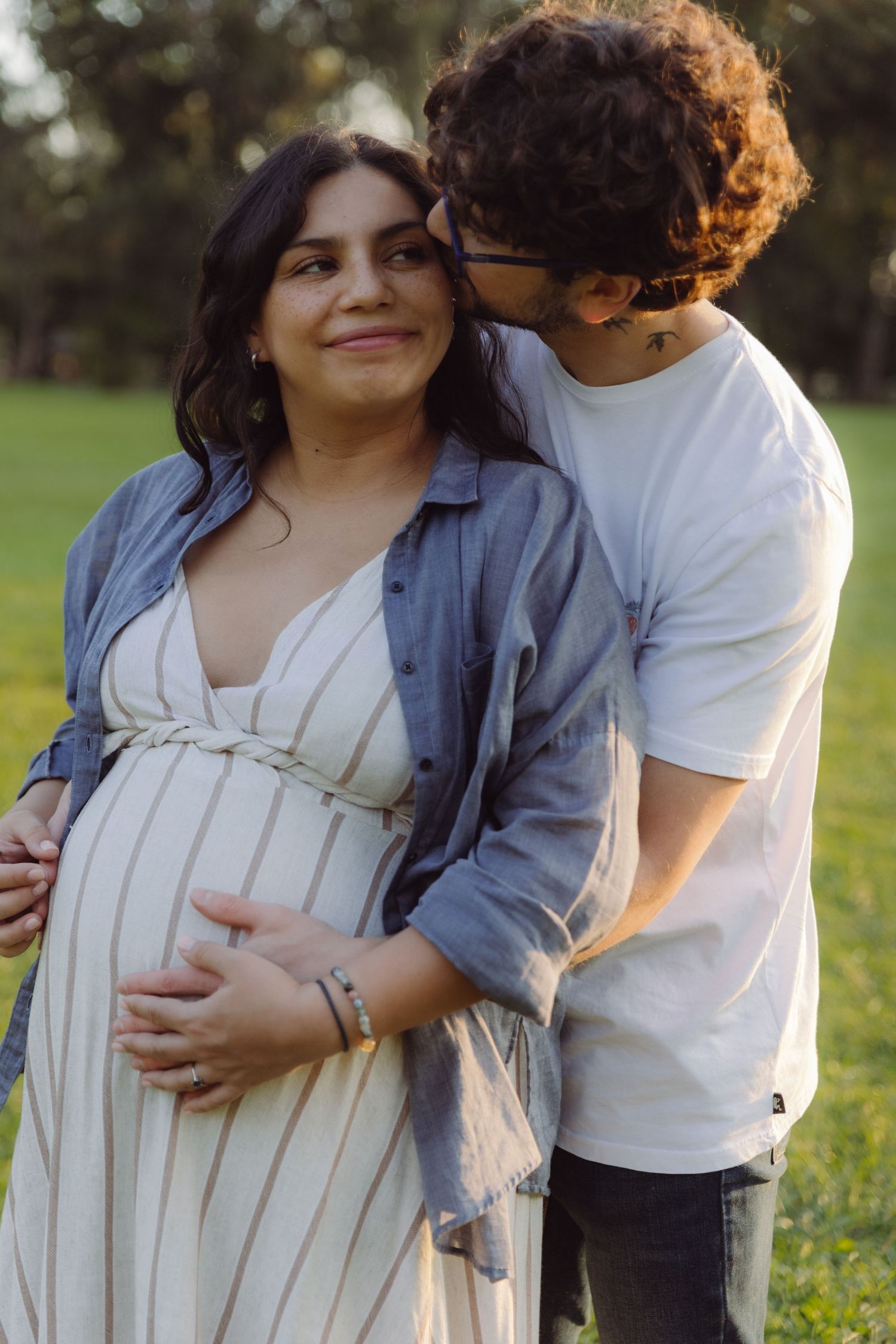Man kissing pregnant woman's cheek while embracing her from behind in sunny park setting.