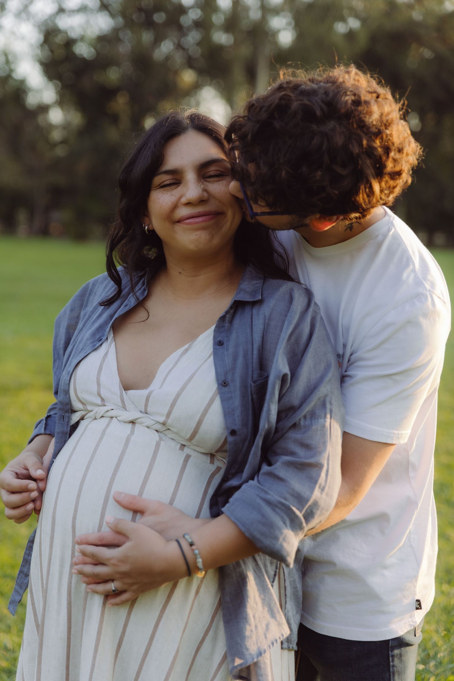 Happy pregnant woman smiling as partner kisses her cheek while cradling her baby bump outdoors.