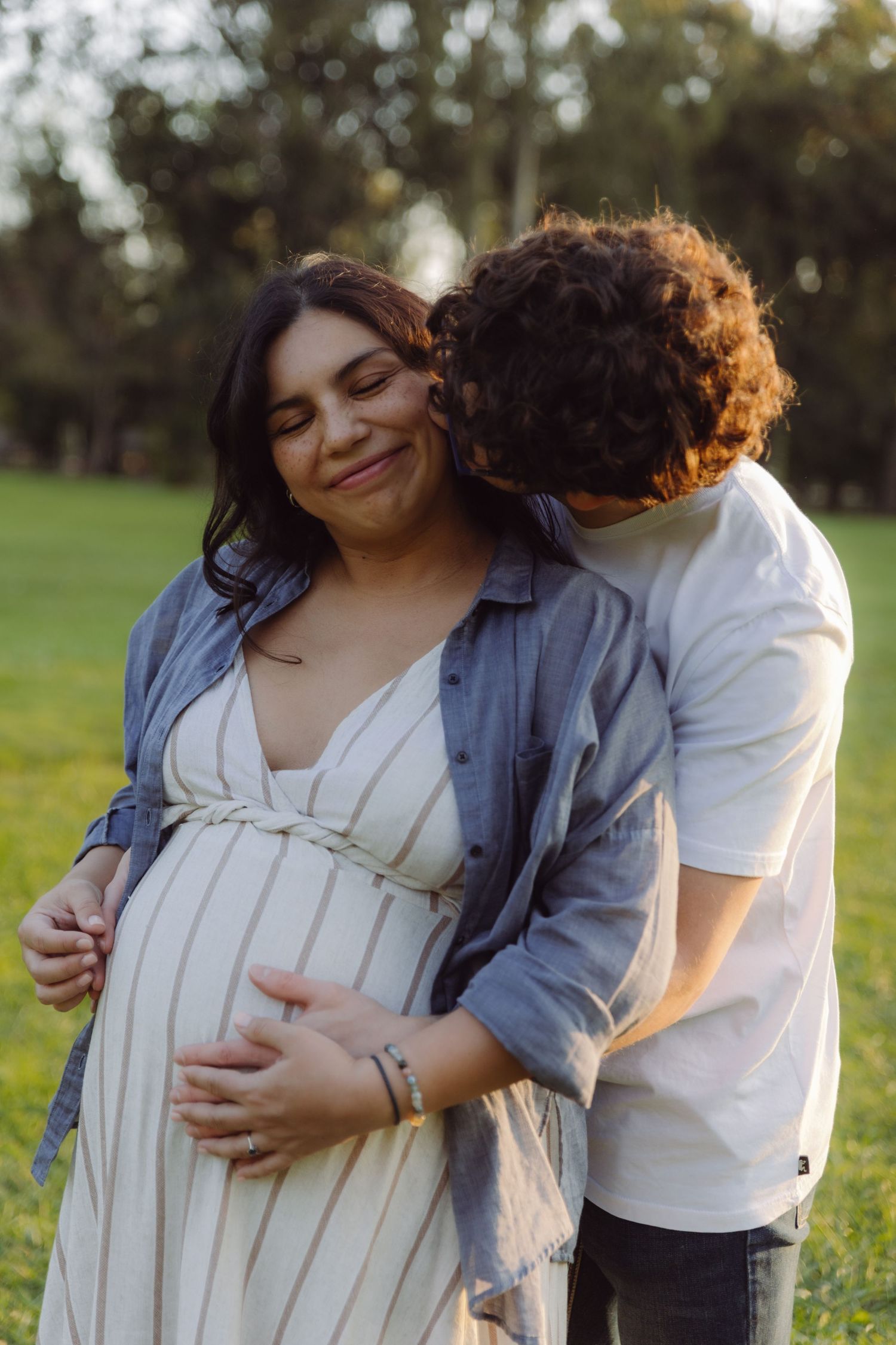 Couple sharing tender moment outdoors, man kissing pregnant woman's cheek while holding her bump.