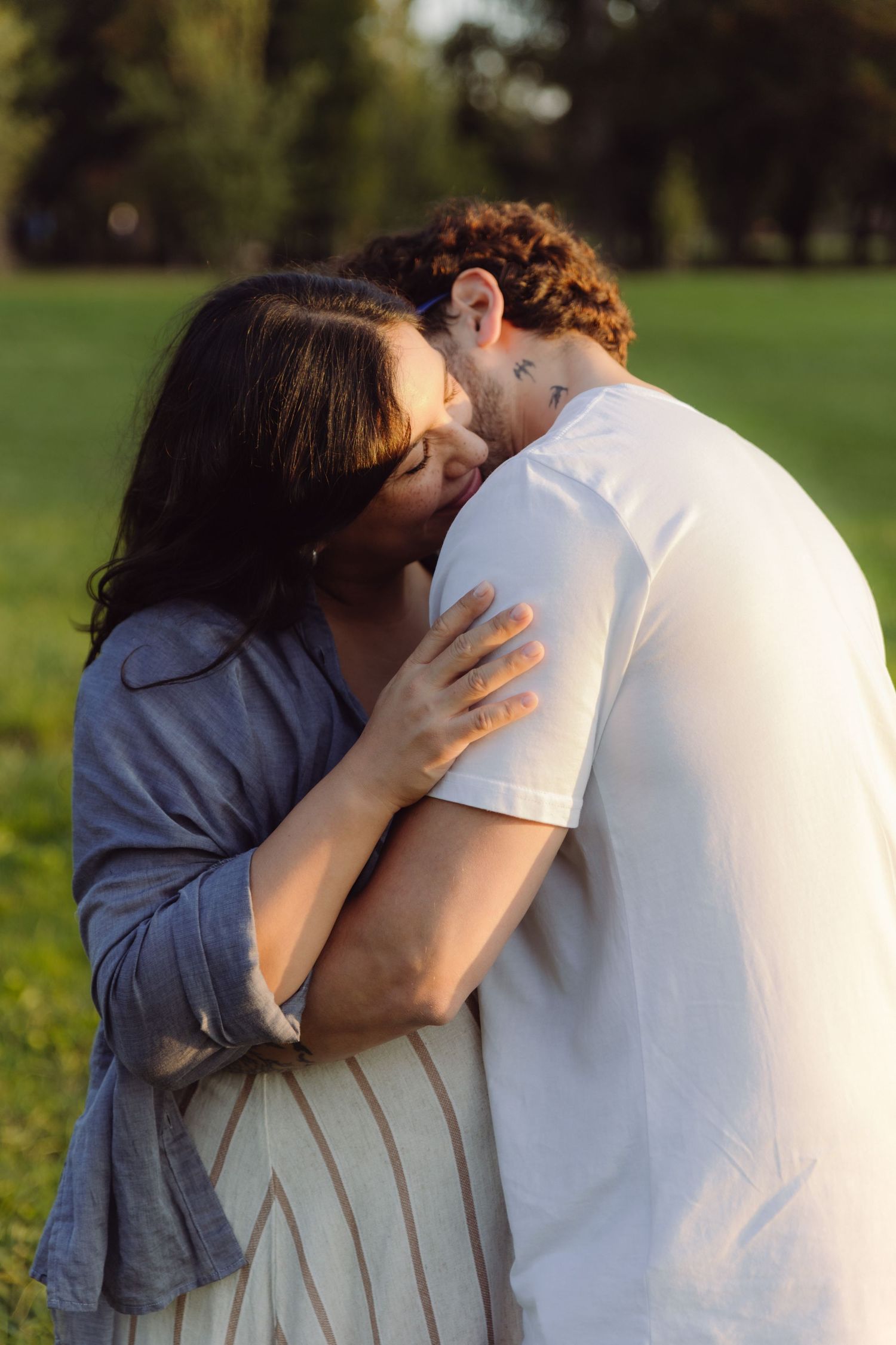 Pregnant couple sharing a kiss while embracing in a sunlit green park at golden hour.