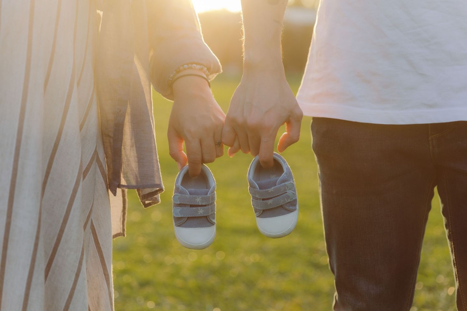 Couple holding hands and tiny baby shoes together outdoors in golden sunset light, announcing pregnancy.