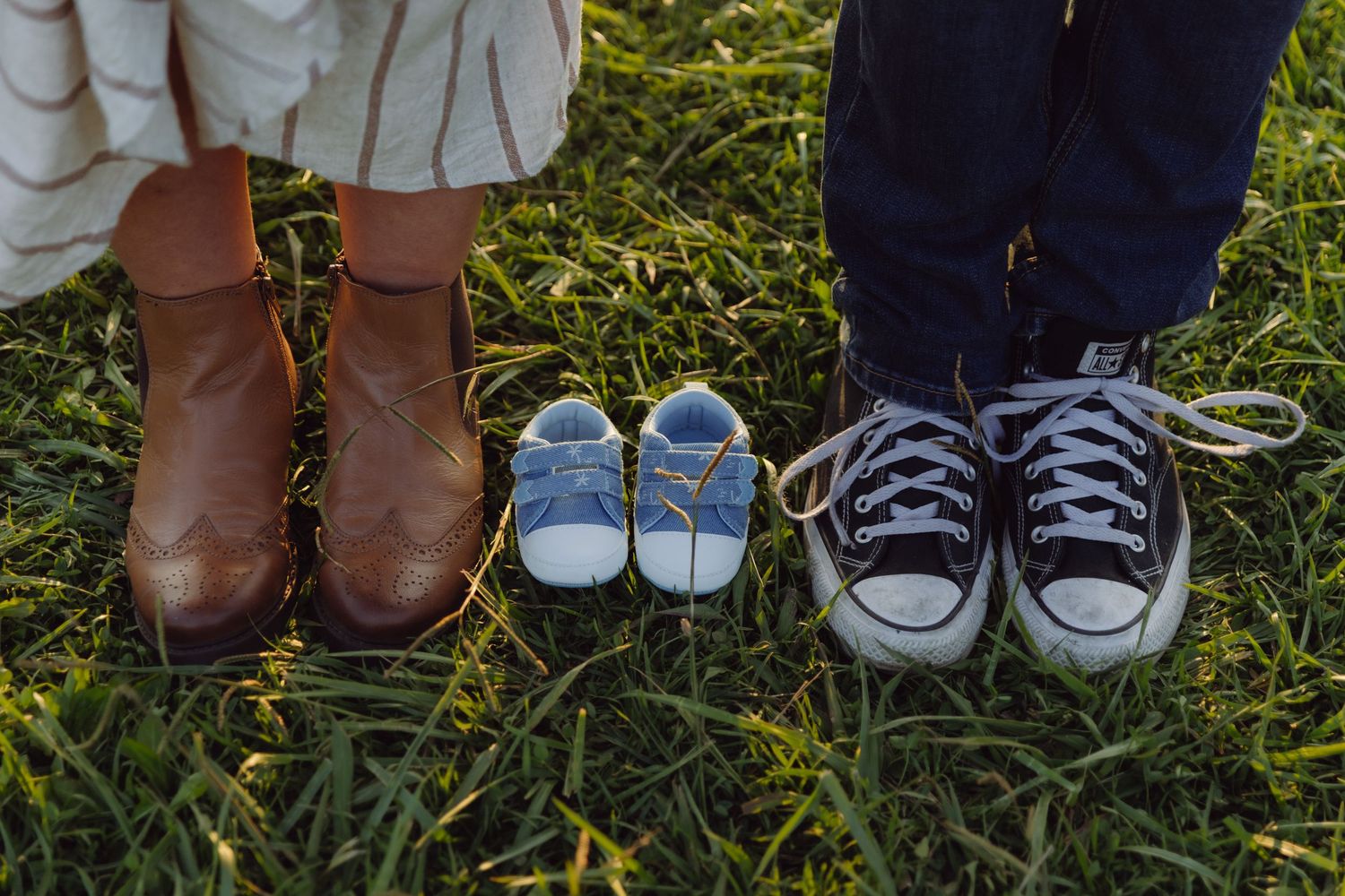 Two pairs of adult shoes with tiny blue baby sneakers between them on green grass.