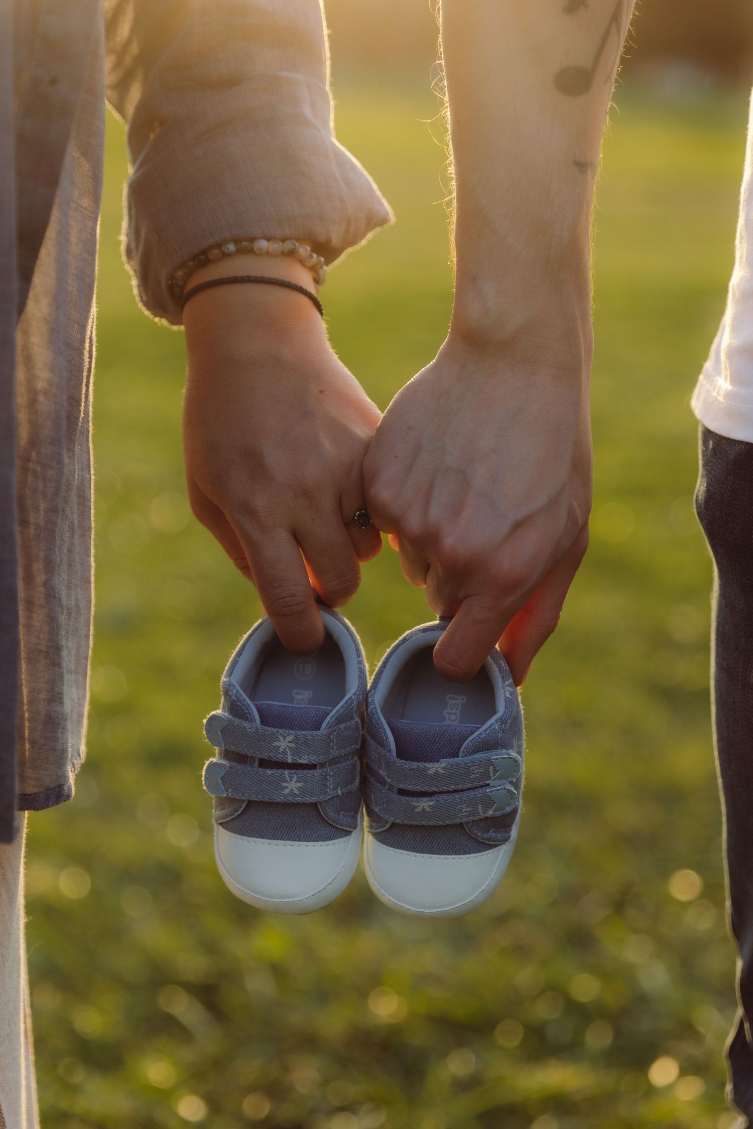 Couple holding tiny baby sneakers together at golden sunset, announcing pregnancy in a park.
