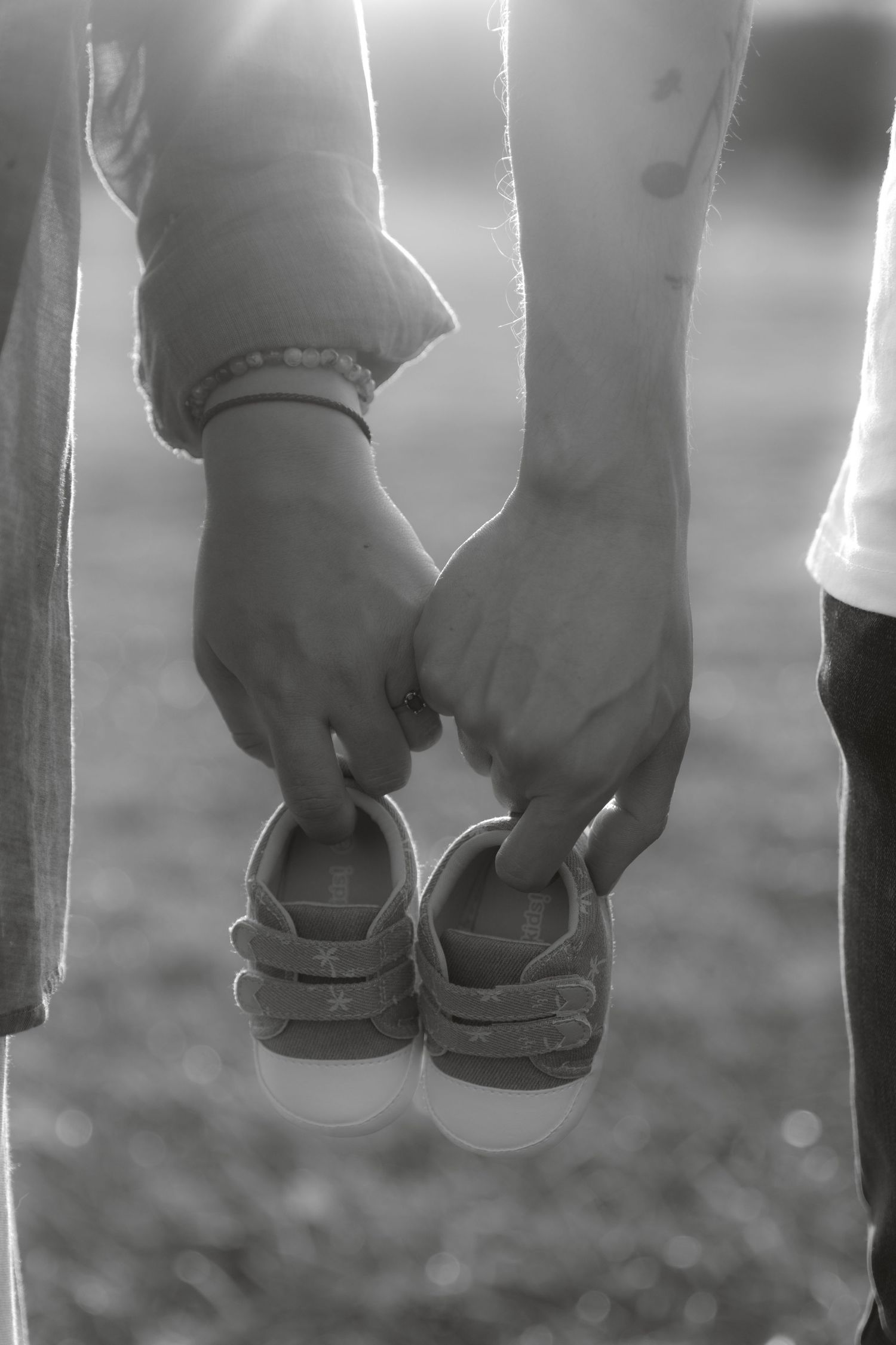 Black and white photo of expectant couple holding tiny baby shoes together in sunlight.