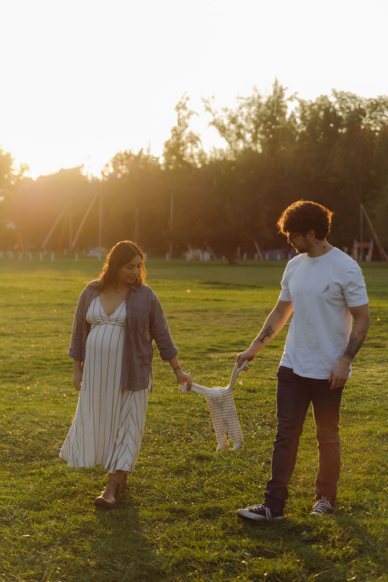 Expecting couple holding baby clothes at golden sunset in open green field.
