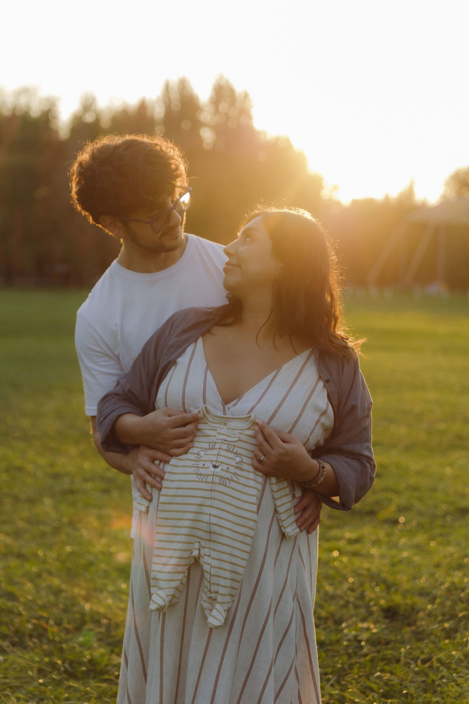 Couple standing in golden sunset light, man embracing pregnant woman holding baby clothing outdoors.