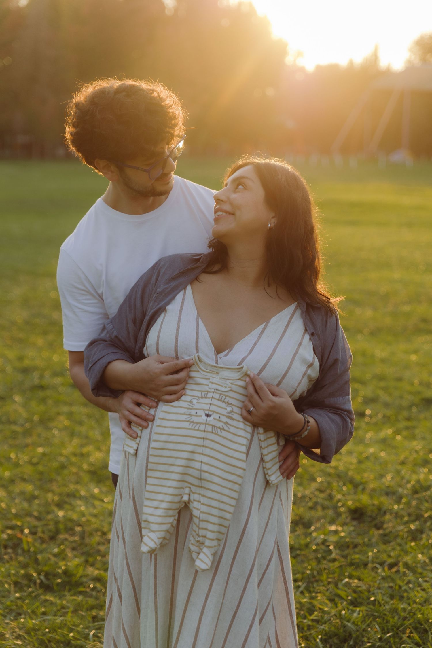 Man embracing pregnant woman holding baby outfit at warm golden sunset outdoors.