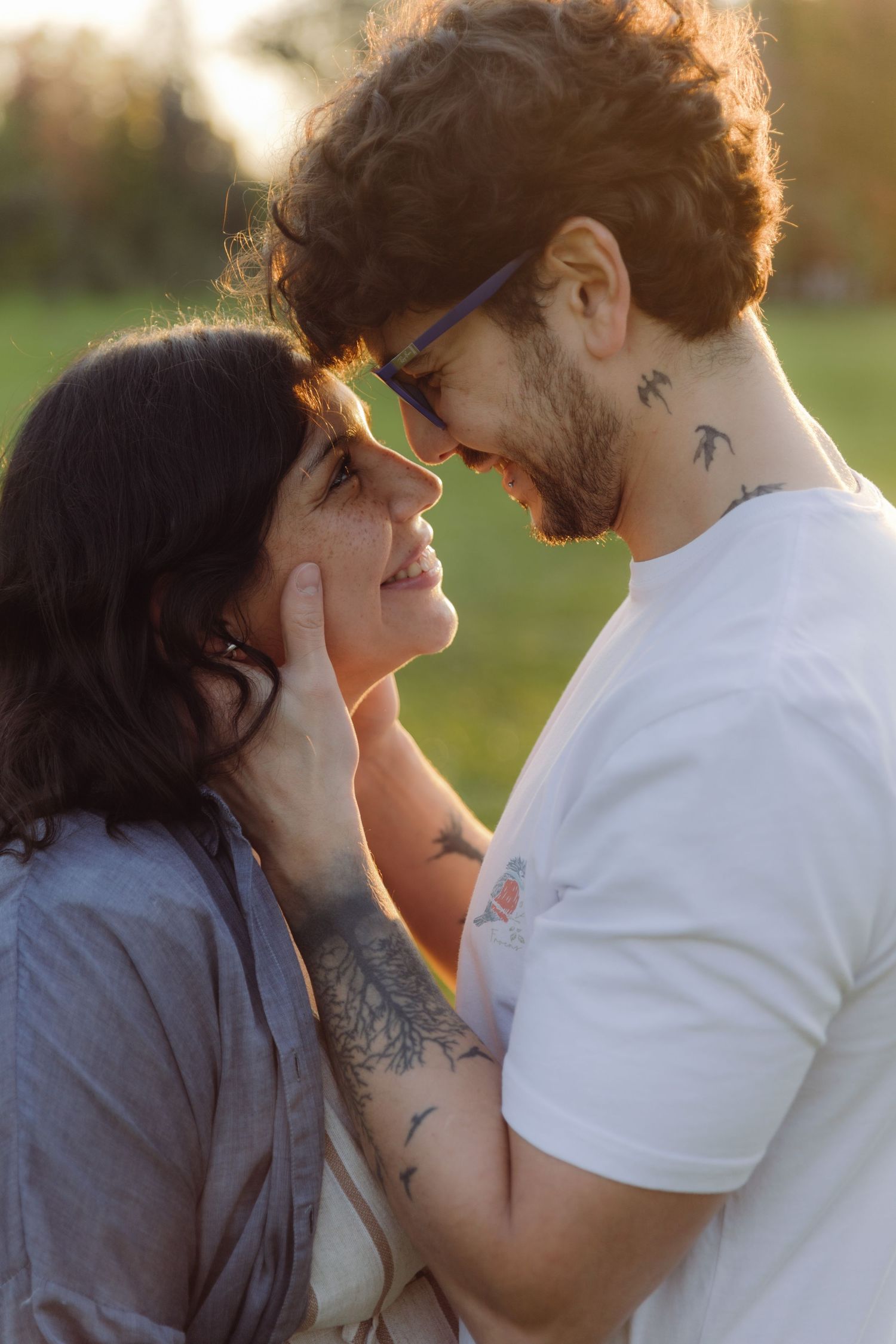 Close-up of tattooed couple gazing into each other's eyes outdoors, man tenderly cupping woman's face at sunset.