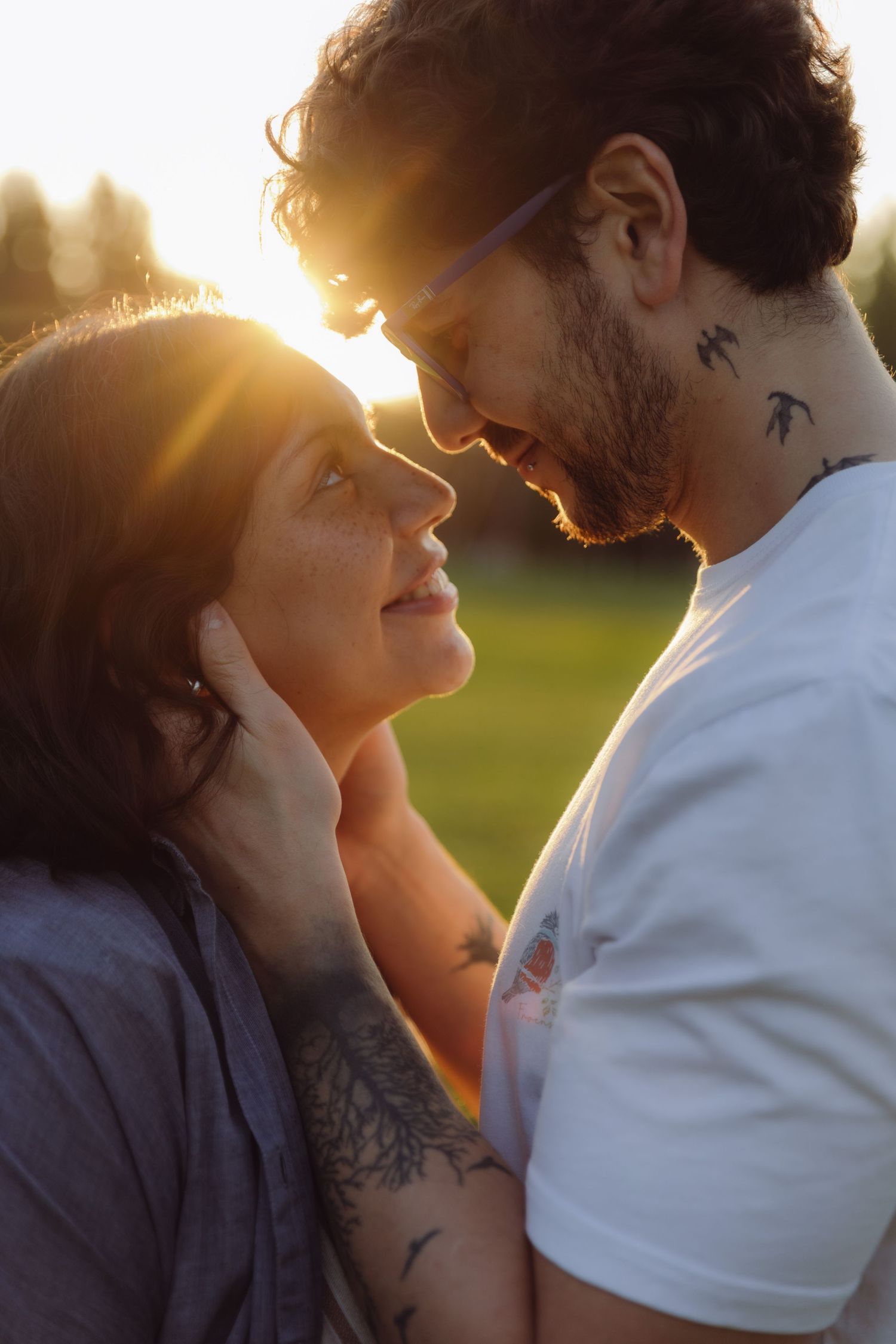 Couple with tattoos sharing a romantic moment at sunset, faces close together in a golden backlit park.