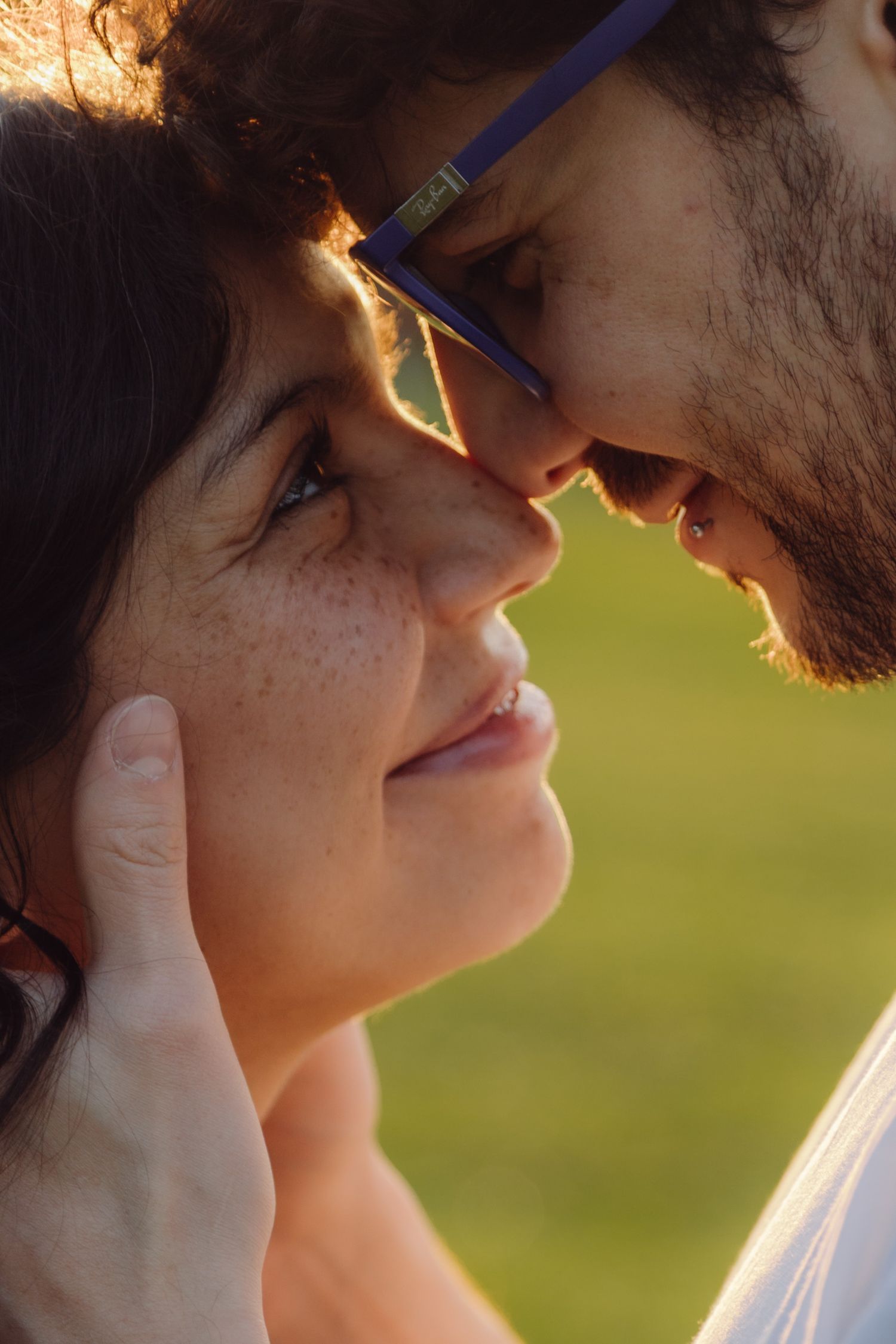 Close-up of a romantic couple almost kissing outdoors, woman with freckles and man with blue glasses in warm sunlight.