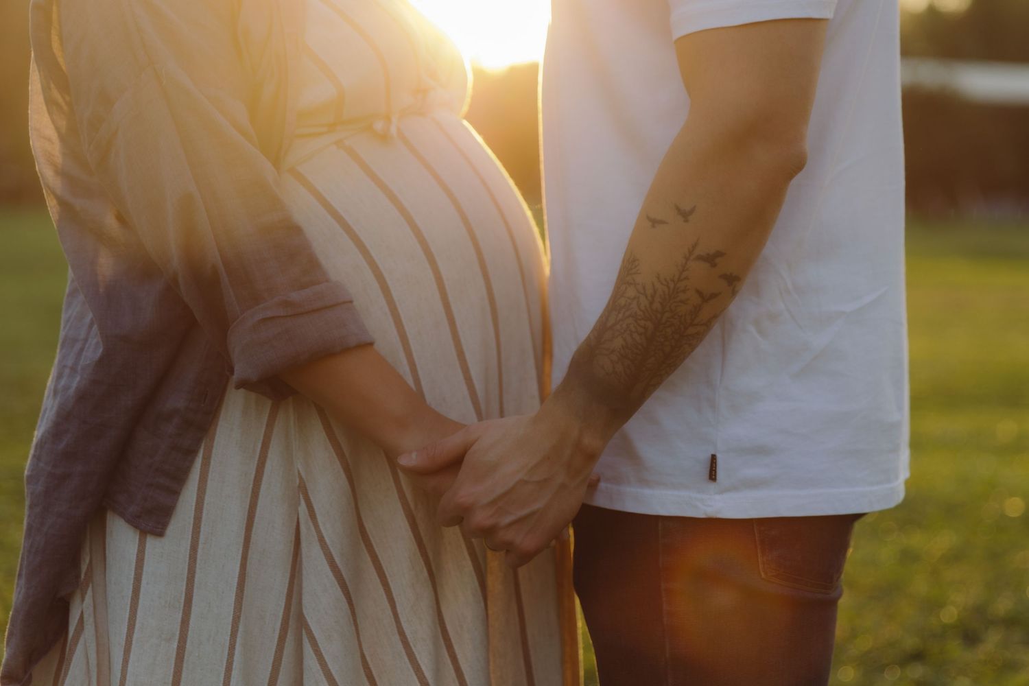 A pregnant couple holds hands outdoors, bathed in warm golden sunset light.