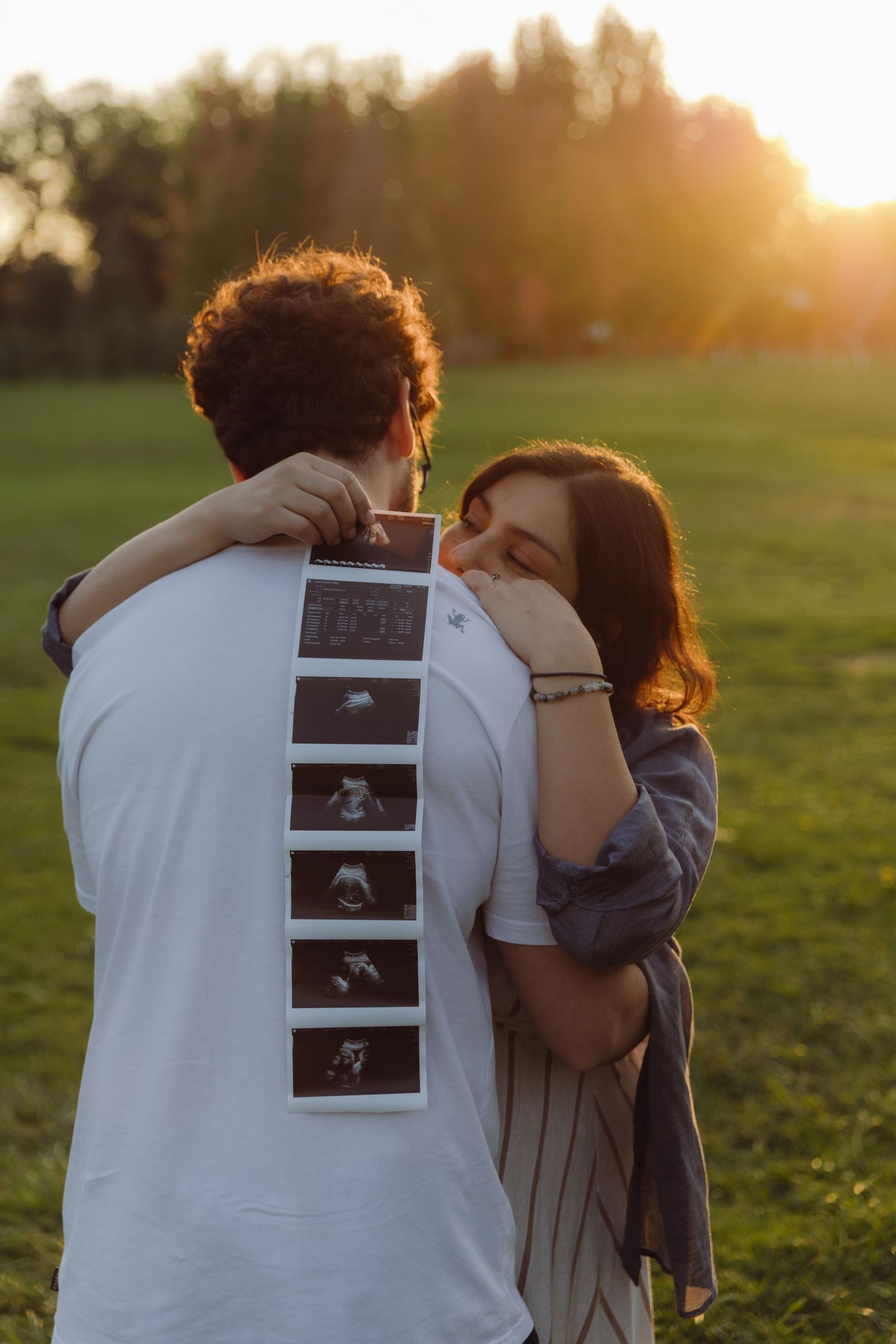Pregnant woman hugging partner from behind showing ultrasound photos at golden hour in park.