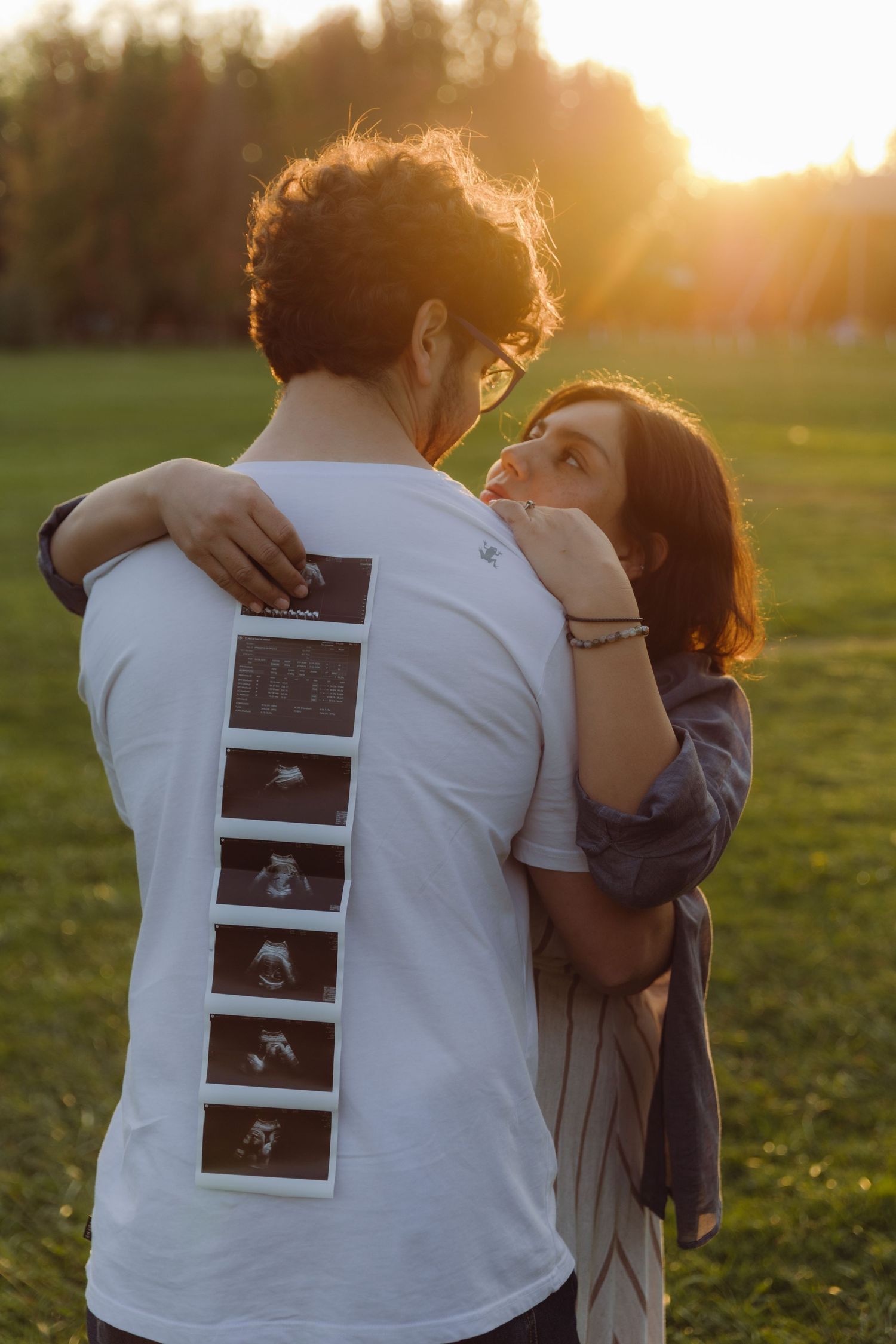 Couple embracing at sunset in park, woman hugging man from behind showing ultrasound photos on his back.