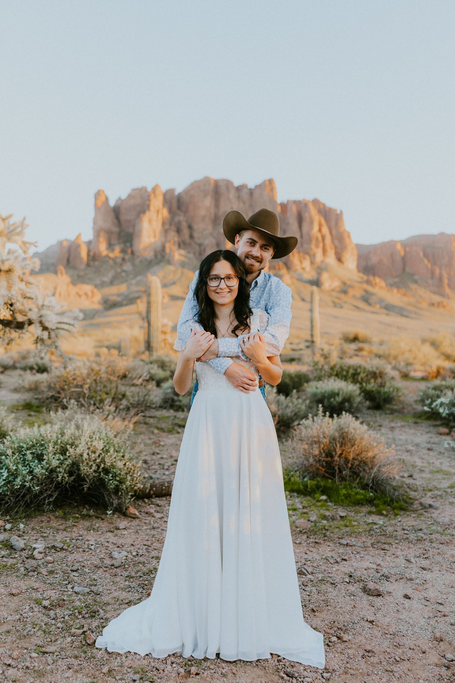 Couple embracing in desert landscape with Superstition Mountains at golden hour, woman in white gown, man in cowboy hat.