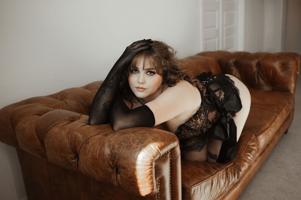 A woman in black lace lingerie and stockings poses on a tufted brown leather sofa.