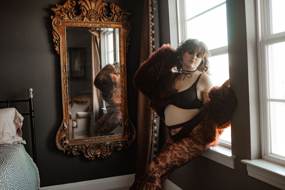 Tattooed woman in black lingerie posing by a window next to an ornate gold mirror in a dark bedroom.