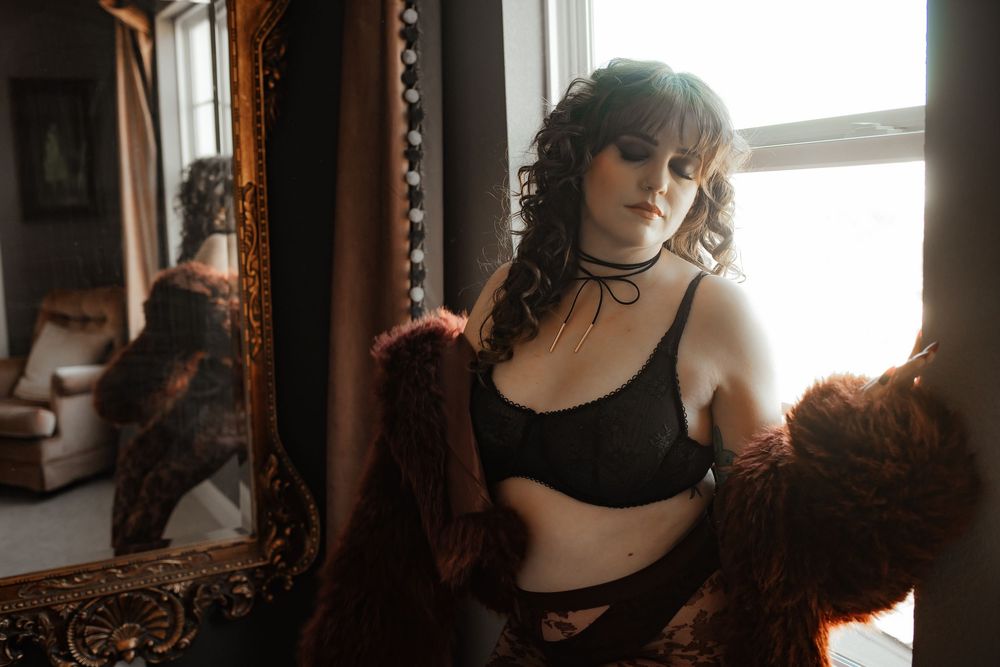 Woman in black lingerie holding a fur coat beside an ornate gold mirror in a softly lit room.
