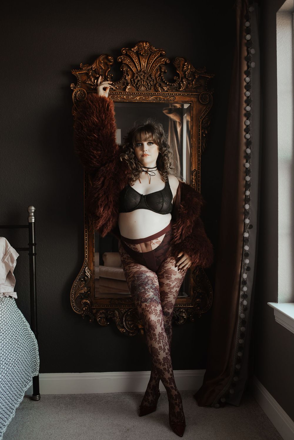 Woman in black lingerie and patterned tights poses before ornate gold mirror in dark moody bedroom.