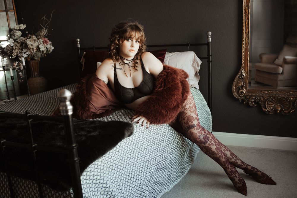 Woman in black lingerie and floral tights poses elegantly on a bed in a moody, dark-toned boudoir setting.