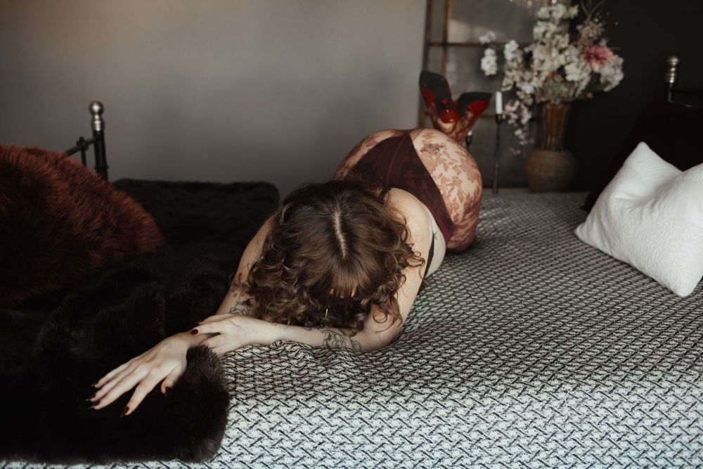A woman in dark lingerie lying curled on a bed with gray bedding, fur accents, and flowers in the background.