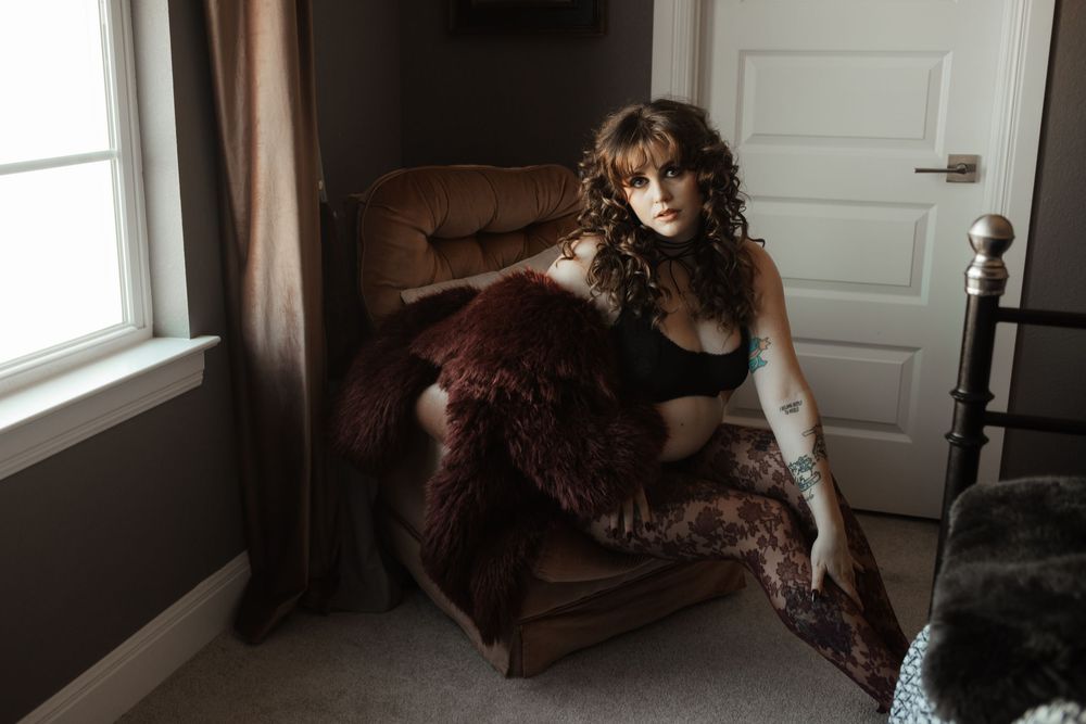 A woman with curly hair sits in a velvet chair wearing black lingerie, draped in a dark fur wrap near a window.