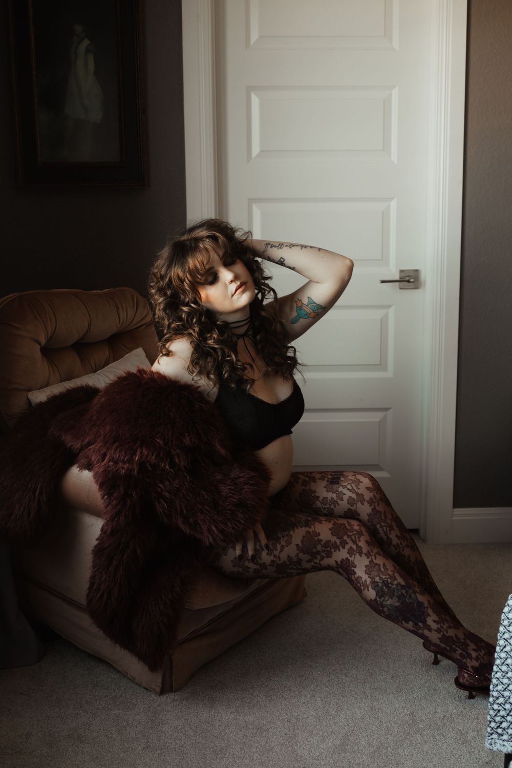 Woman in black lingerie and lace tights poses elegantly on floor beside a velvet chair in moody bedroom setting.