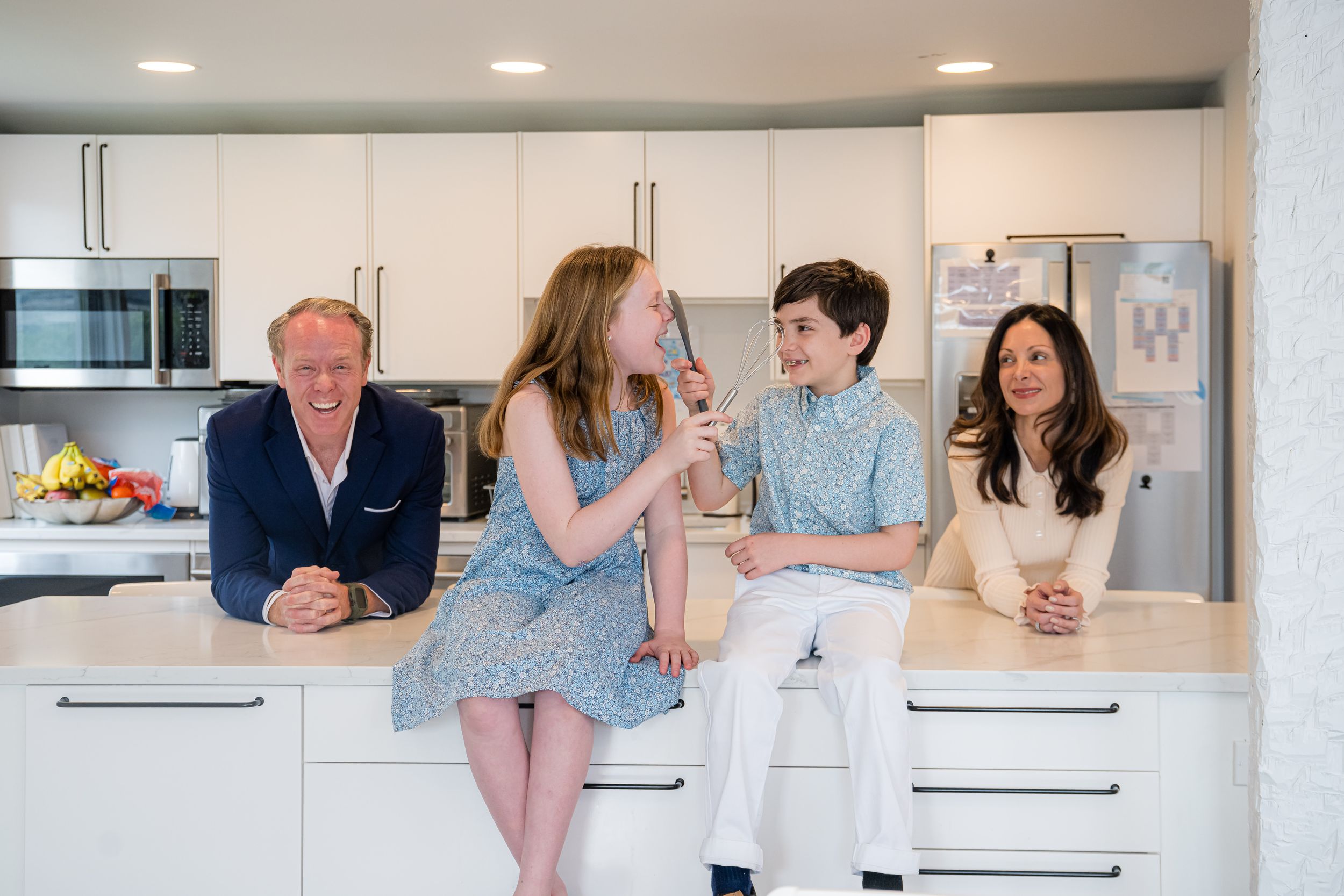 Family of four laughing together in a modern white kitchen, sitting at a bright countertop in casual elegant attire.