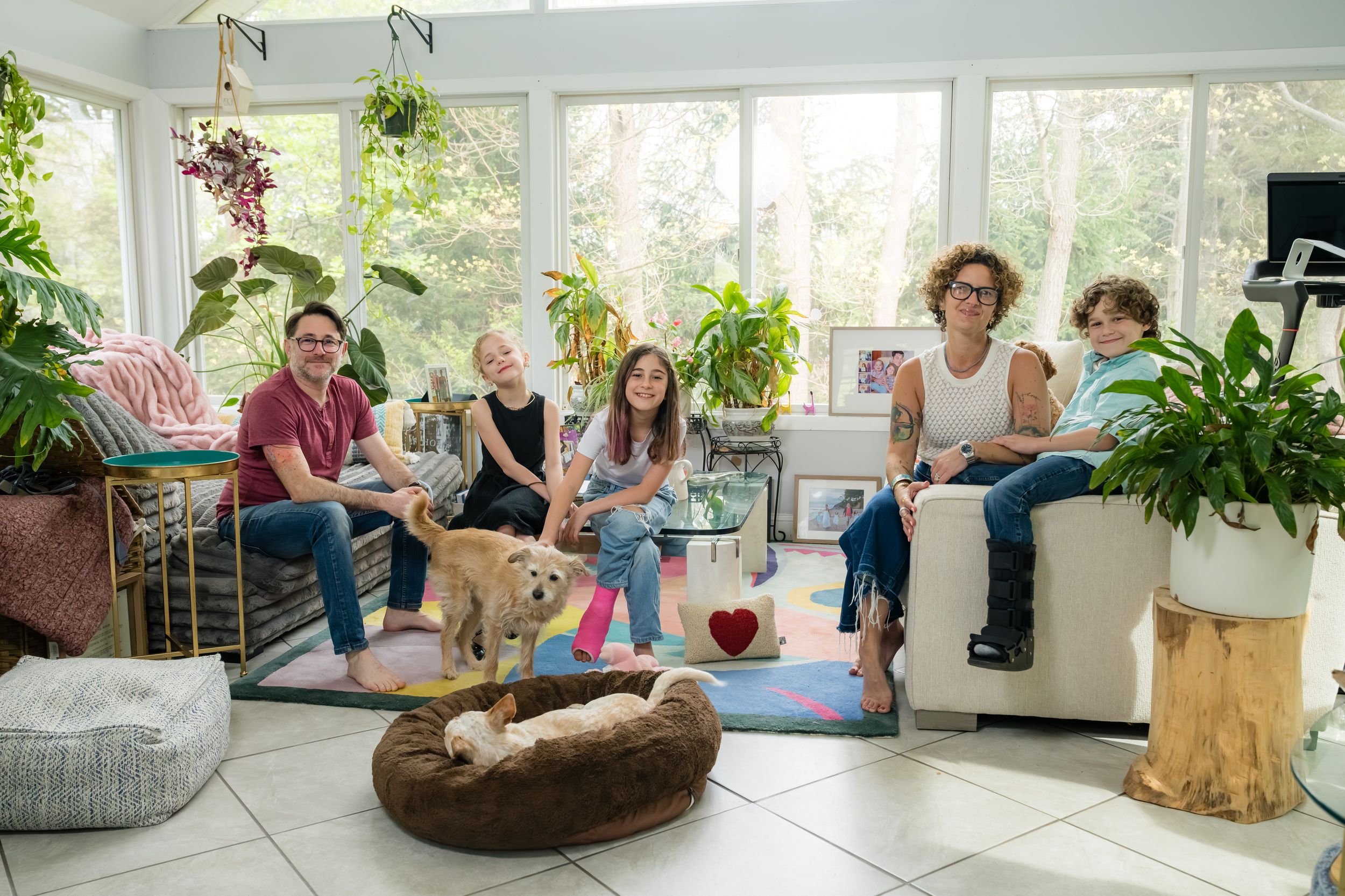Family with dog relaxing in bright sunroom surrounded by lush green plants on tiled floor.