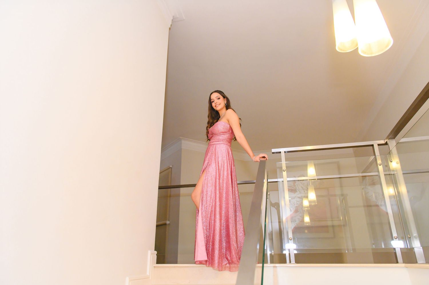 Woman in elegant pink satin gown standing gracefully on modern white staircase with glass railing.