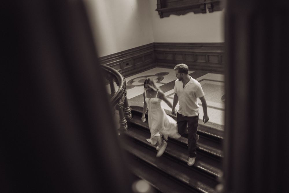 Black and white photo of a couple holding hands walking up grand staircase in elegant building.