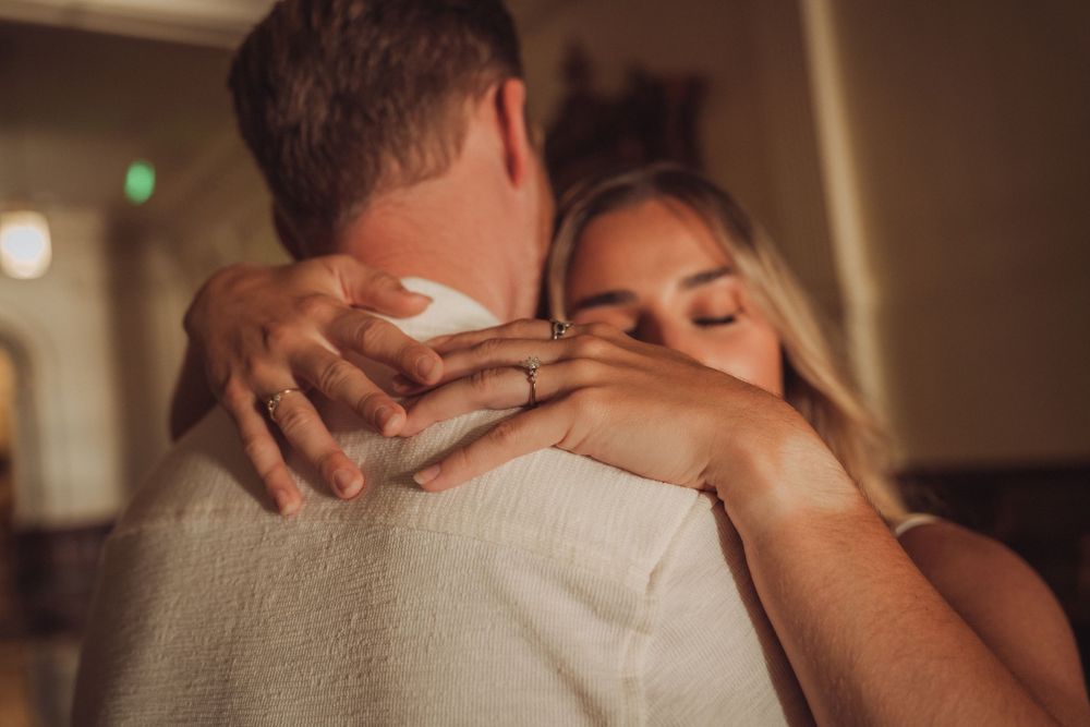 Couple sharing an intimate embrace, woman with blonde hair closing her eyes while hugging man in white shirt at indoor venue.