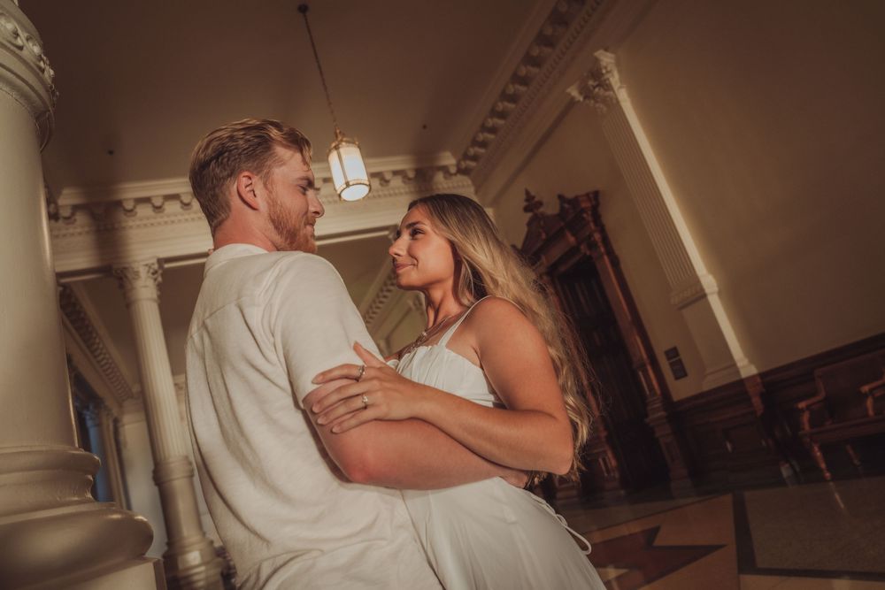 Couple in white attire sharing a romantic moment between grand columns in an elegant venue.