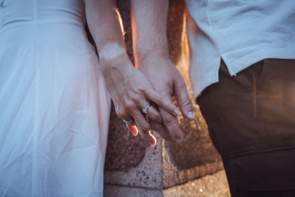Close-up of a couple holding hands, woman wearing an engagement ring, sitting together in soft warm light.