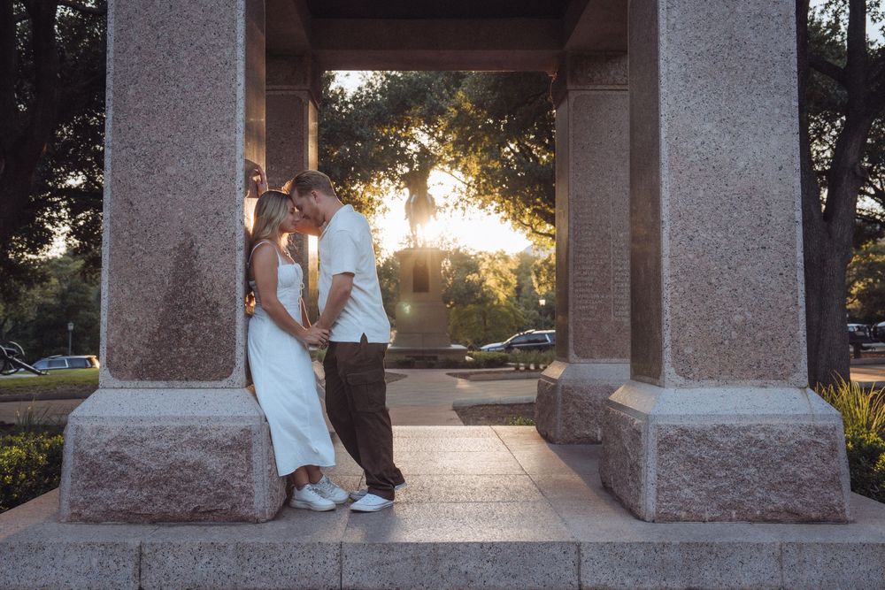 Couple sharing a romantic moment kissing between granite columns at sunset in a park.