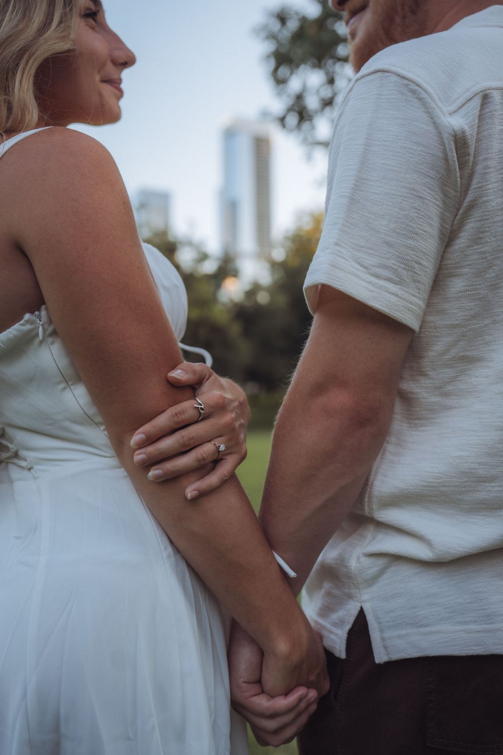 Couple holding hands outdoors in park, woman in white wedding dress with engagement ring, city skyline in background.