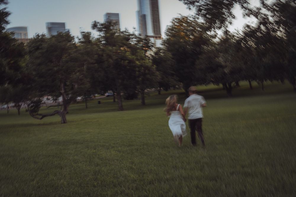 A couple runs hand-in-hand through a lush green park with city skyscrapers visible in the background at dusk.