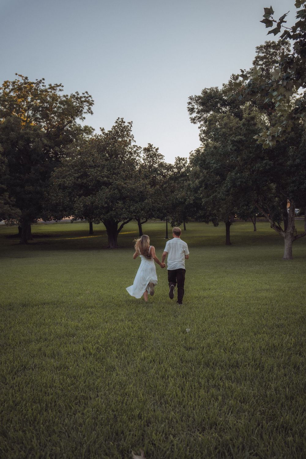 Two children holding hands running through a lush green park surrounded by trees at dusk.