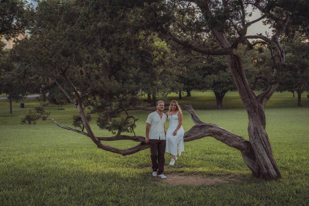 Couple in white outfits posing under a curved tree in a lush green park at dusk.