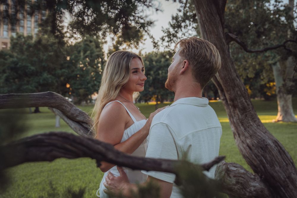 A couple shares a romantic moment among trees in a lush green park at golden hour.