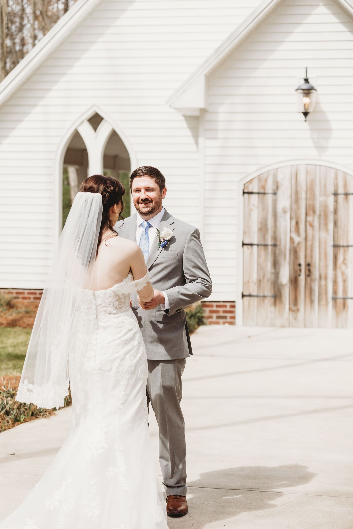 Groom in gray suit smiles at bride in white lace gown during first look outside a white chapel.