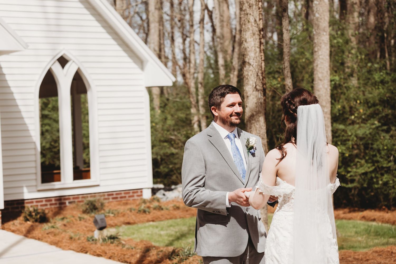 Groom in gray suit shares first look moment with bride in white gown and veil outside white chapel.