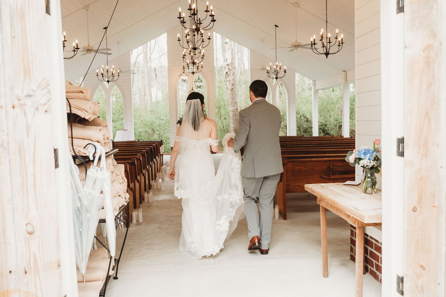 Bride and groom walking down the aisle of a bright white chapel with chandeliers and wooden pews.