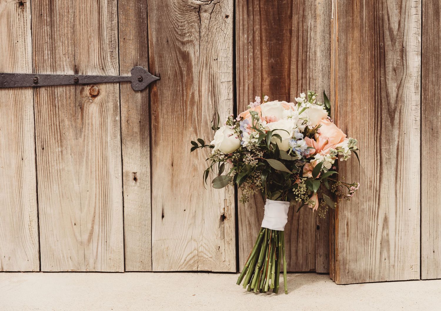A rustic wedding bouquet with peach roses and white flowers leans against a weathered wooden barn door.