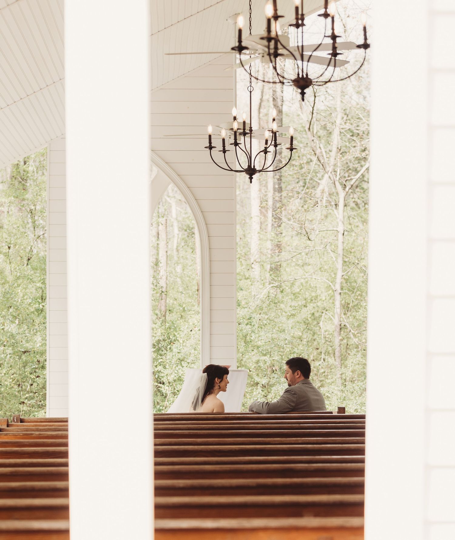 Bride and groom sit together in wooden pews of a white chapel with arched windows and elegant chandeliers.