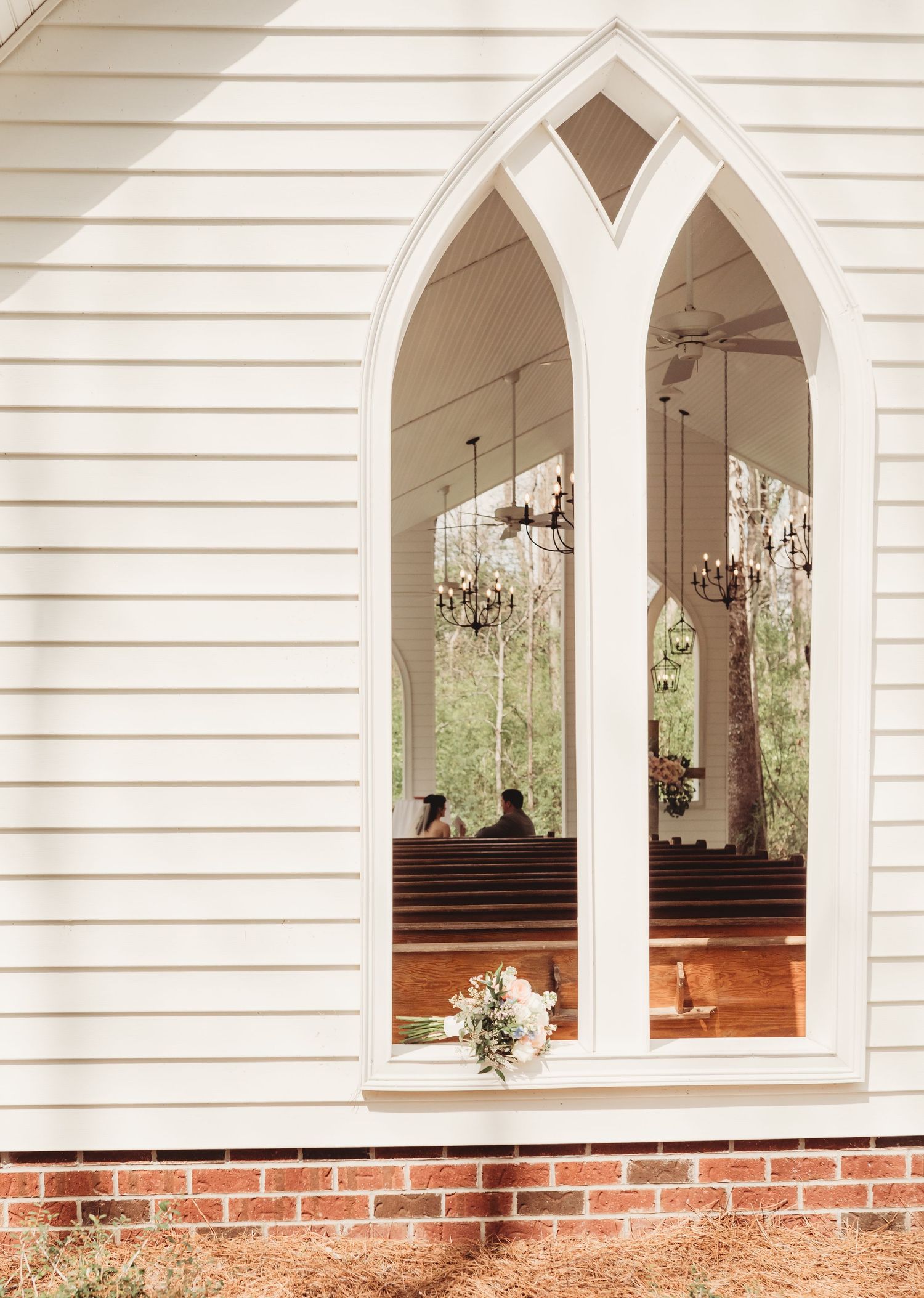 Gothic arched window of a white chapel with brick foundation, reflecting chandeliers and wedding guests inside.