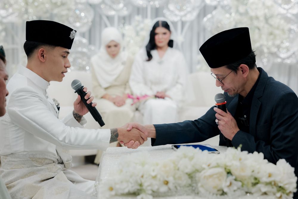 Two men in traditional Malay attire shake hands at a wedding ceremony with guests in white seated behind.