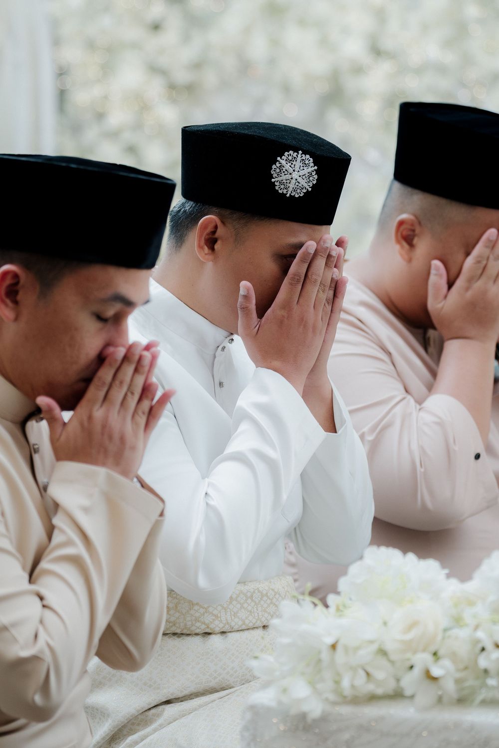 Three Muslim men in traditional Baju Melayu and songkok hats praying with hands raised during a religious ceremony.