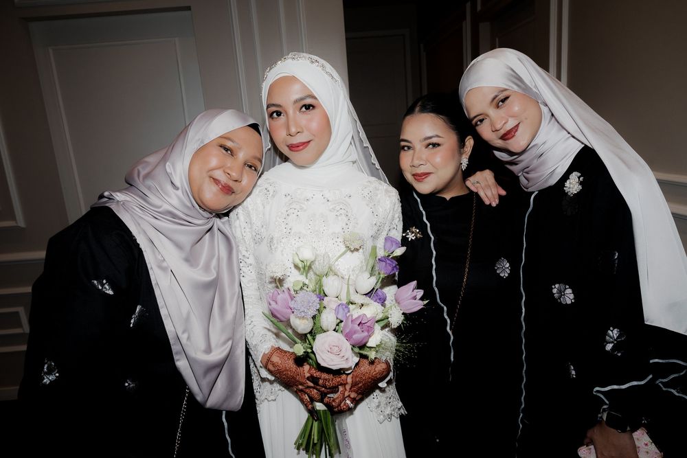 Four women in hijabs pose together, with a bride in white holding a bouquet of purple and white flowers.